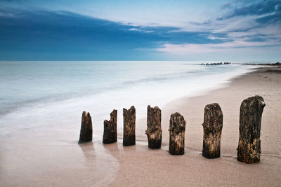 Weathered wooden posts in sand at beach against sky