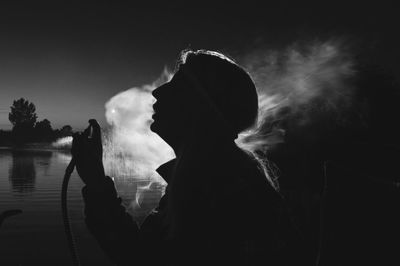 Side view of silhouette young woman smoking by lake at night