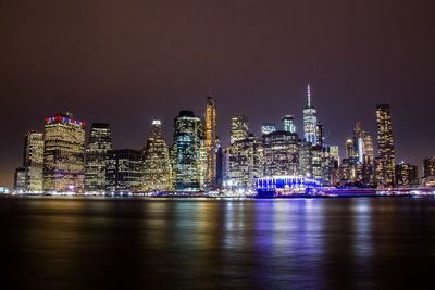 Illuminated modern buildings by river against sky at night