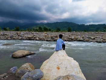 Rear view of man sitting on rock at river against sky
