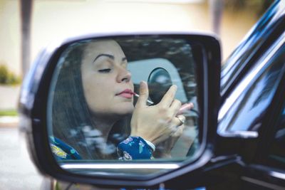 Woman applying lip gloss reflecting on side-view mirror of car