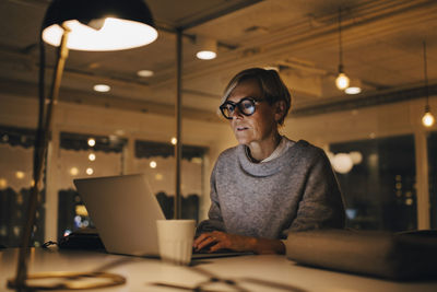 Confident mature female business professional using laptop while sitting at desk working late in office