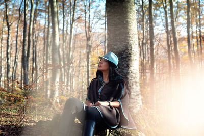 Woman looking away sitting by tree trunk in forest