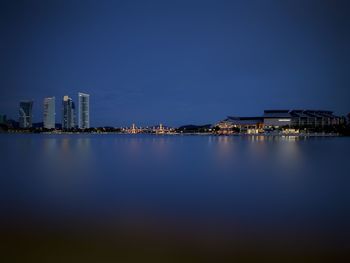 Illuminated buildings by sea against blue sky at night