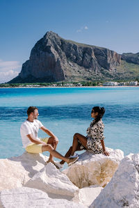 Woman sitting on rock by sea against mountain