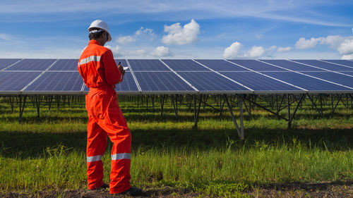 Rear view of man examining solar panels while standing on grassy field against blue sky