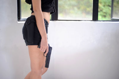 Midsection of woman holding gun while standing against wall