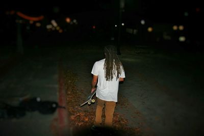 Rear view of man with long hair holding skateboard on footpath at night