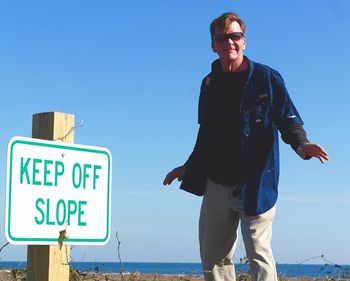 Portrait of mature man standing by information sign at beach against clear blue sky during sunny day