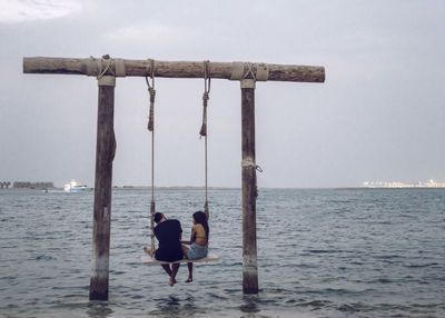 Rear view of couple sitting on rope swing over sea against sky