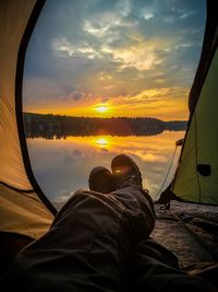 Low section of person resting in tent by river against sky during sunset