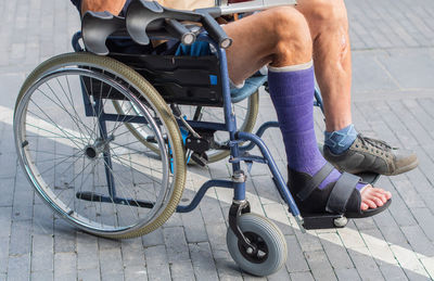 Sitting man in wheelchair with purple plaster cast on leg and crutches nearby on tiled terrace