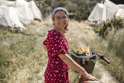 Happy woman with vegetables in bicycle basket standing in farm