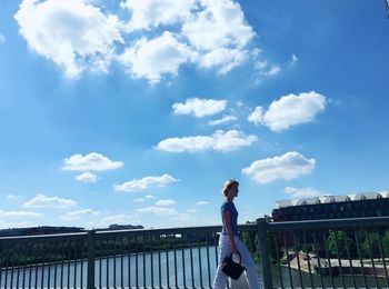 Woman walking by railing of bridge against cloudy sky