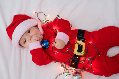 A baby boy dressed in a santa suit lies on the bed and plays with a christmas garland