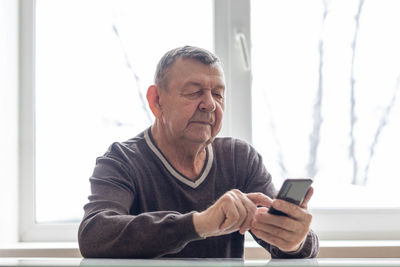 Elderly man dials number in smartphone from home. old senior sends sms on mobile phone