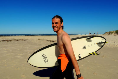 Portrait of shirtless man standing on beach against clear blue sky