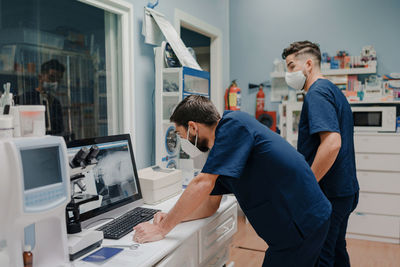 Anonymous young male doctors in respiratory masks and uniforms working on desktop computer in clinic