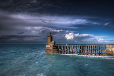 Lighthouse by pier in front of sea against cloudy sky