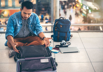 Man looking at open suitcase while sitting on bridge in city