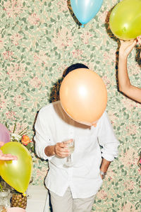 Obscured face of young man holding drink while standing by woman with balloons against wallpaper at home during dinner p