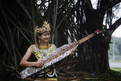 Smiling woman playing traditional musical instrument against trees