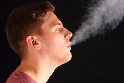 Close-up of young man exhaling smoke against black background