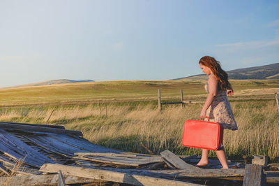 Full length of young woman holding red suitcase while walking on abandoned wood against sky
