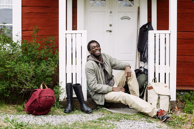 Portrait of happy young man with backpacks and boots sitting outside cottage