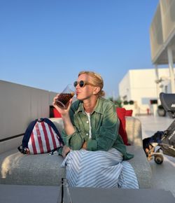 A young woman with light hair, putting a backpack, is resting on a sofa, drinking a soft drink