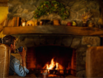 Man having drink while sitting by fireplace at home