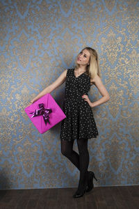 Thoughtful young woman holding pink gift box against patterned wall