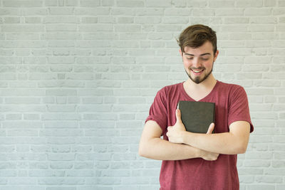 Smiling young man holding bible while standing against brick wall