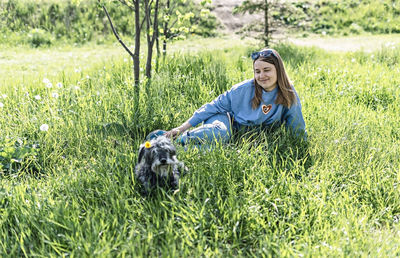 Pretty young woman resting with fluffy dog on green grass among dandelions in summer, pet love, care