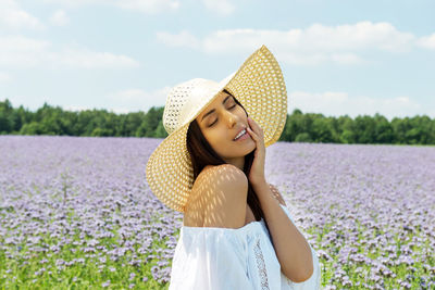 Smiling young woman with eyes closed wearing hat while standing amidst plants against sky