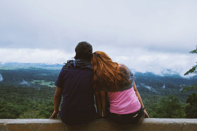 Rear view of couple sitting on retaining wall against sky