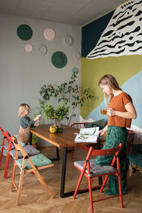 Mother reads magazine standing at table, child waters plants on table