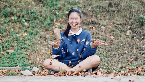 Portrait of smiling young woman throwing leaves while sitting on field in park