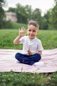 Portrait of a four year old boy in a white shirt on a field in summer sit on cover and show five