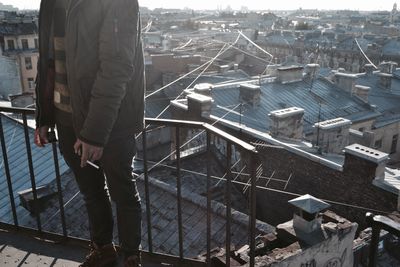 Low section of man standing with cigarette by railing against cityscape