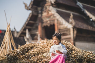 Girl sitting on haystack against cottage outdoors