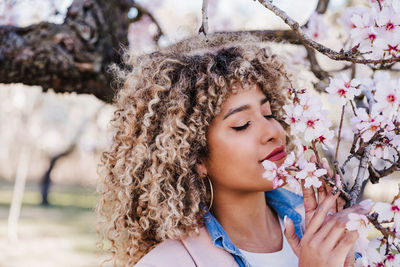 Portrait of beautiful hispanic woman with afro hair in spring smelling pink blossom flowers. nature