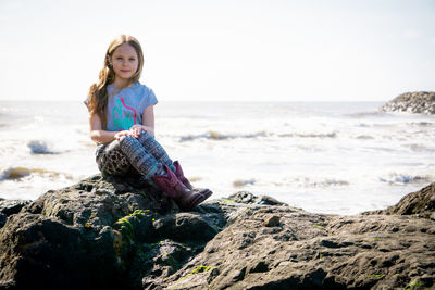 Full length of girl sitting on rock at beach against sky