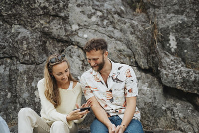 Smiling woman sharing smart phone with male friend sitting in front of rocks