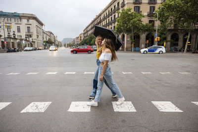 Happy young woman holding umbrella with friend crossing street in city