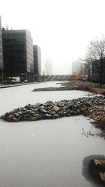 Frozen river by buildings in city against clear sky