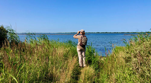 Rear view of woman standing at green bank of lake, watching with binoculars