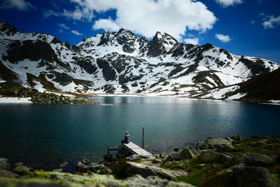 Woman sitting with scenic view of snowcapped mountains by lake against sky 