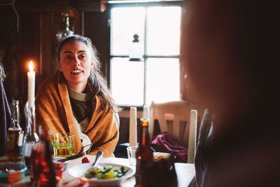 Young woman wearing blanket looking away while sitting at table against window in log cabin