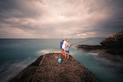 Men fishing while standing on rock in sea against cloudy sky